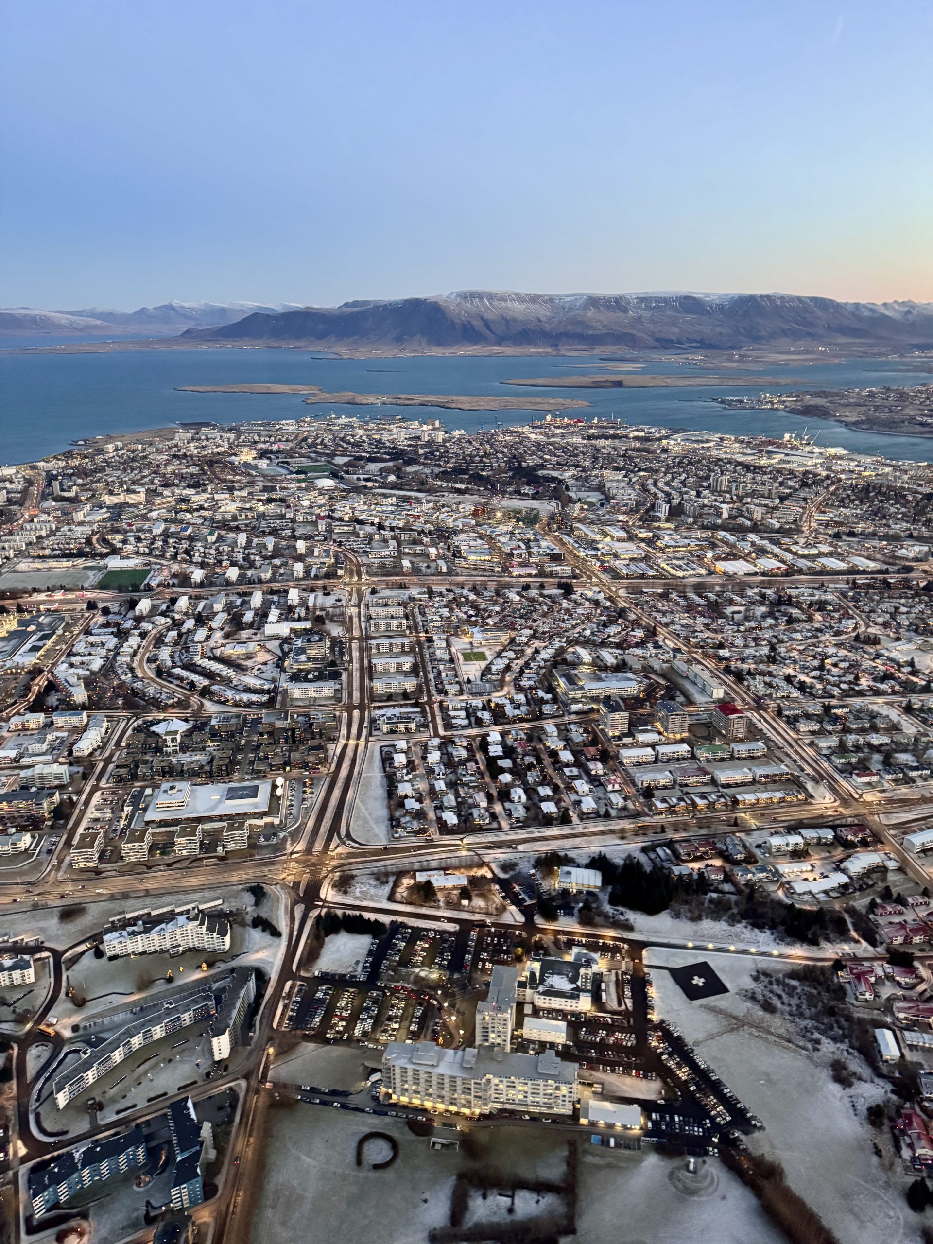 Luxury aerial view of Reykjavík city taken from a private helicopter, showing the coastline, city lights, and surrounding mountains.
