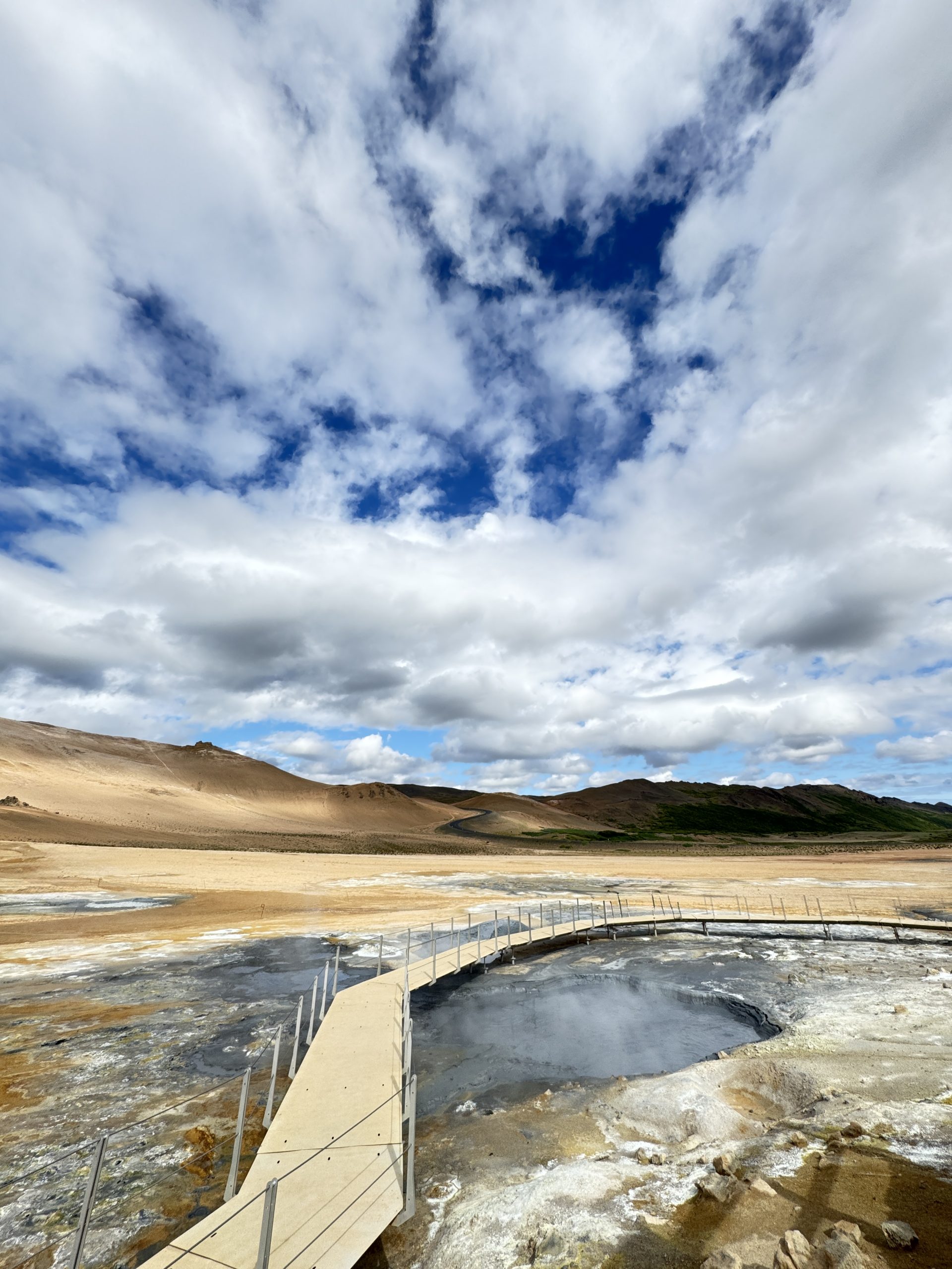 Wooden boardwalk over a steaming geothermal hot spring at Hverir in North Iceland under a cloudy blue sky.